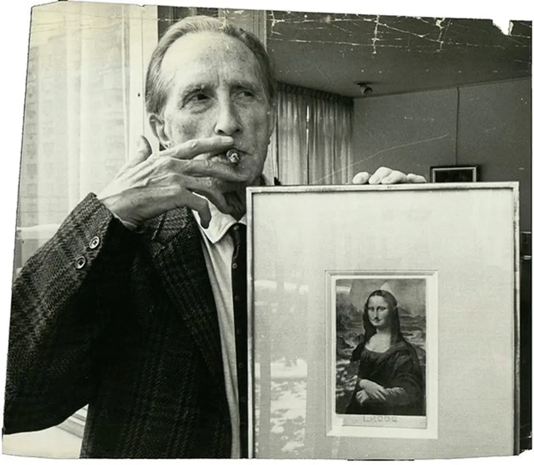Marcel Duchamp, smoking a cigar, holding up a framed copy of L.H.O.O.Q.
