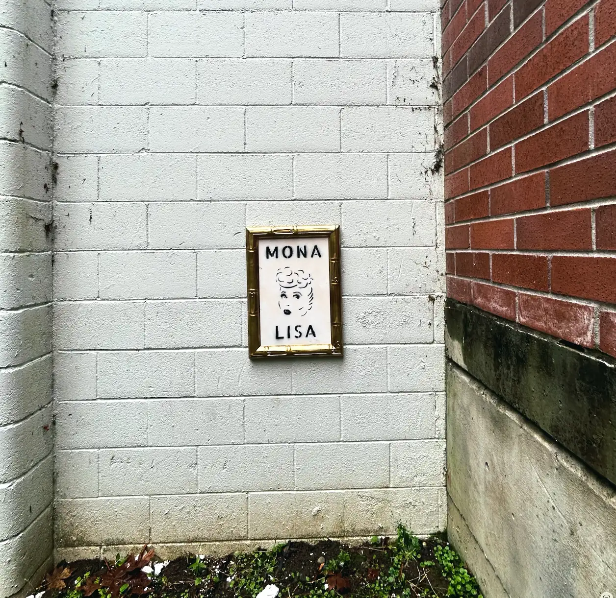norville’s framed Mona Lisa hanging alone on a white cinder-block wall in an alley corner, beside a red brick building