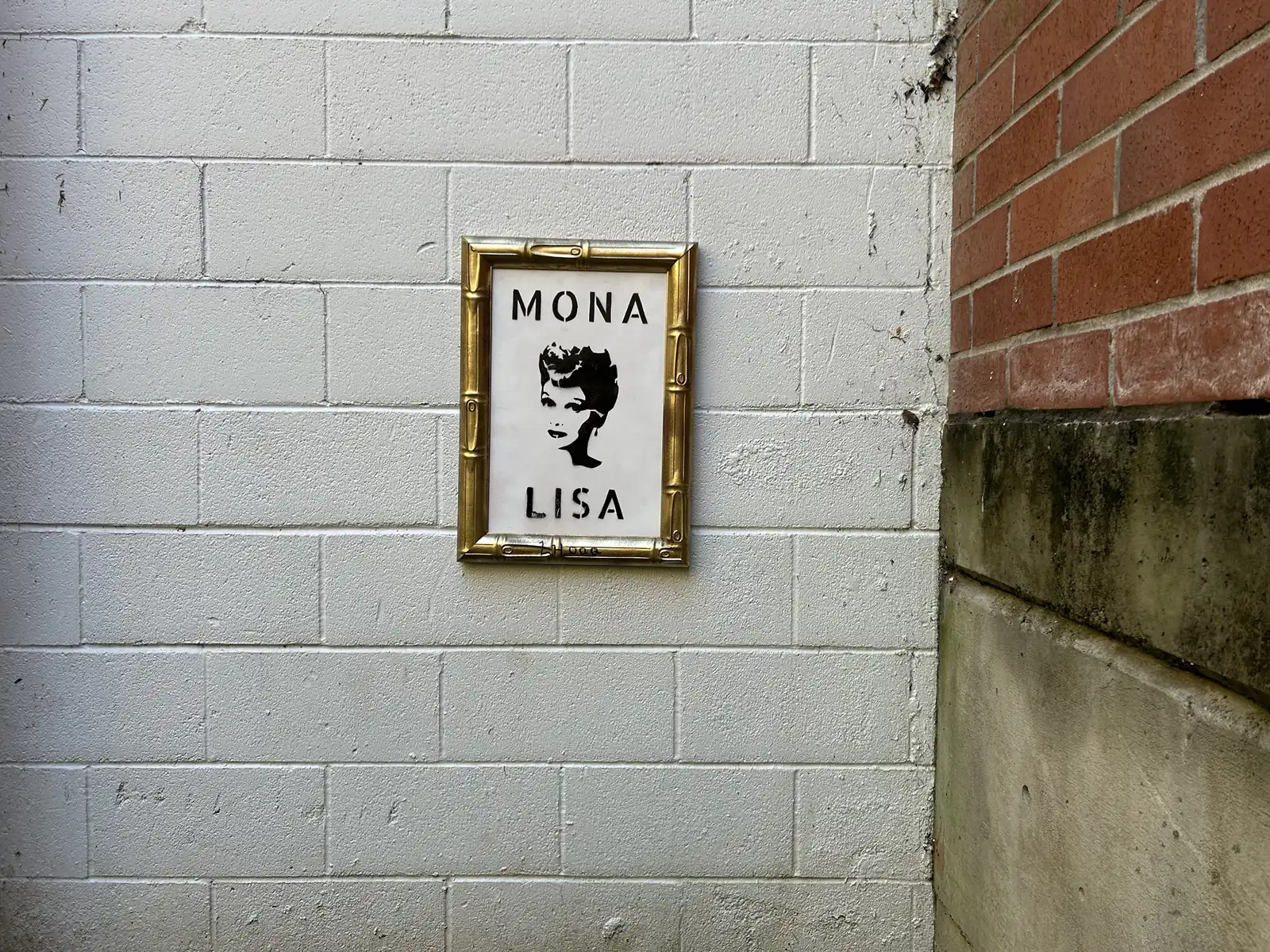 norville’s framed Mona Lisa, a stenciled portrait in a gold bamboo frame, hanging on a white cinder-block wall next to a red brick building